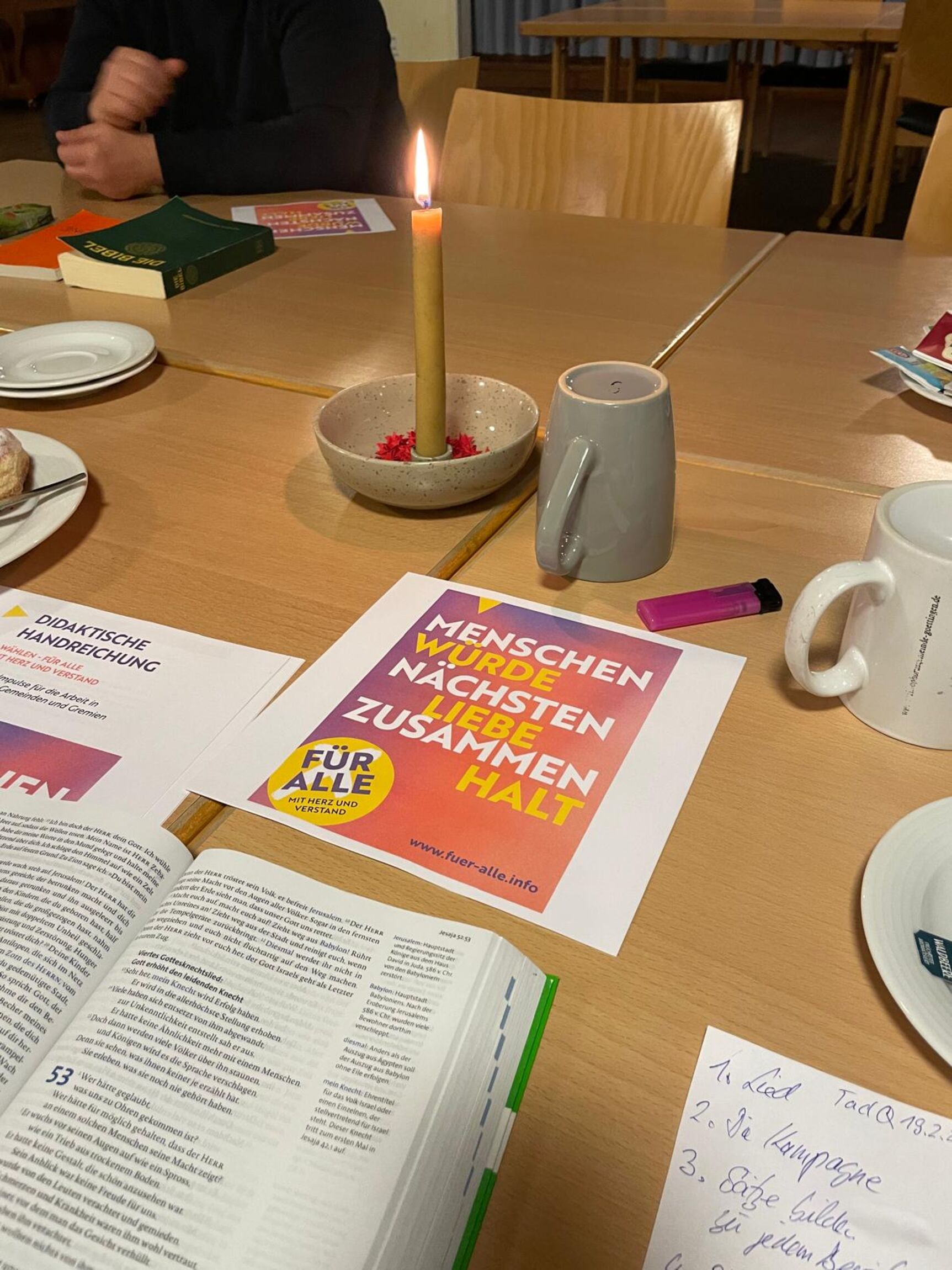 Nahaufnahme Tisch beim Gesprächsabend Treff an der Quelle, im Vordergrund aufgeschlagene Bibel, eine angezündete Kerze, Teebecher, im Hintergrund sitzt jemand mit einer geschlossenen Bibel vor sich
